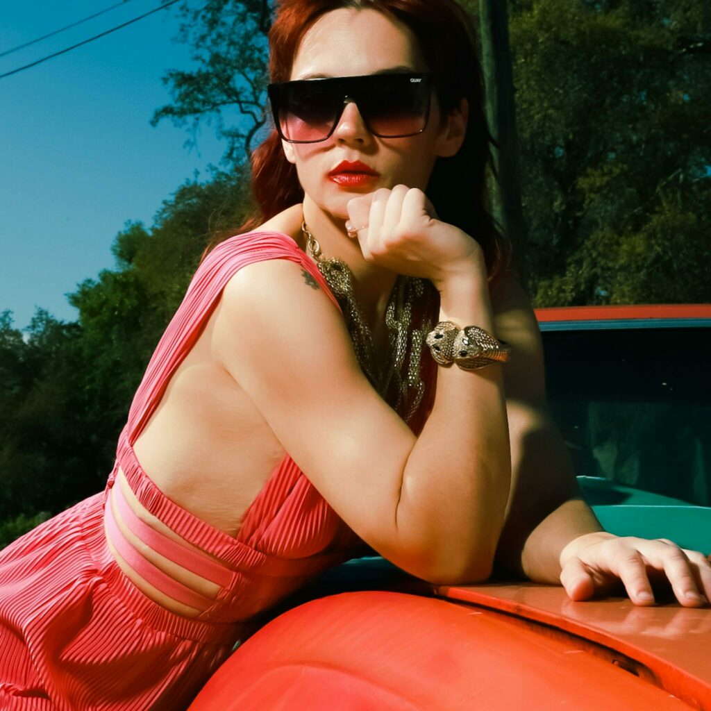 Confident woman in sunglasses poses against a classic car under sunny skies.